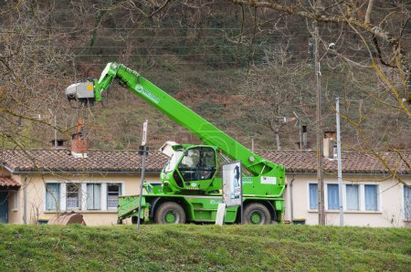 Photo for Les Avalats, France - Jan. 28, 2024 - A green construction telehandler with a hydraulic boom parked on the curbside, in front of a block of countryside houses built on the hillside near a worksite - Royalty Free Image