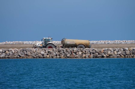 Téléchargez les photos : Port-la-Nouvelle, France - 5 avril 2024 - Un tracteur remorque un réservoir pour pulvérisation de liant hydraulique, sur le projet d'extension du port industriel de Port-la-Nouvelle, sur la mer Méditerranée - en image libre de droit