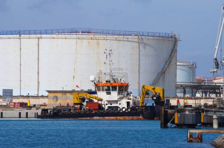 Téléchargez les photos : Port-la-Nouvelle, France - 5 avril 2024 - Un bateau industriel avec des grues amarrées par des réservoirs de stockage circulaires dans le terminal pétrolier du port commercial de Port-La-Nouvelle, sur la côte méditerranéenne - en image libre de droit