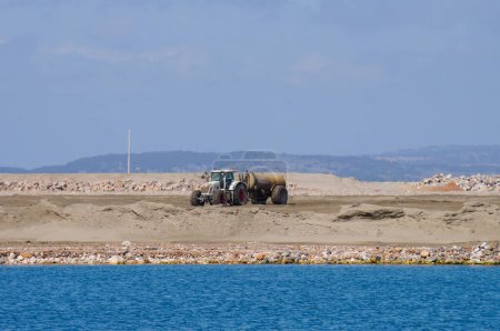 Foto de Port-la-Nouvelle, Francia - 5 de abril de 2024 - Un tractor remolcando un tanque para la propagación de aglutinantes hidráulicos, en el proyecto de extensión del puerto industrial de Port-la-Nouvelle, en el mar Mediterráneo - Imagen libre de derechos