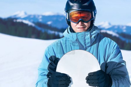 Photo for Portrait of man at the ski resort on the background of mountains and blue sky,  hold snowboard. Wearing ski glasses in the mountains - Royalty Free Image
