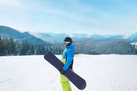 Photo for Snowboarder Of Man At Ski Resort On The Background Blue Sky,  Hold Snowboard. Wearing Ski Glasses. Ski Goggles  With The Reflection Of Snowed Mountains - Royalty Free Image