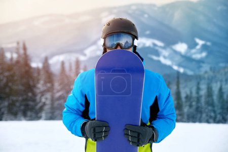 Photo for Portrait Of Man On The Background Of Mountains And Blue Sky, Holding Snowboard And Wearing Ski Glasses. Ski Goggles Of Snowboarder With The Reflection Of Snowed Mountains. Winter Sports - Royalty Free Image