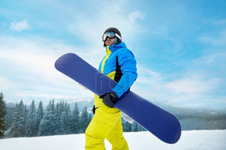Photo for Portrait Of Man On The Background Of Mountains And Blue Sky, Holding Snowboard And Wearing Ski Glasses. Ski Goggles Of Snowboarder With The Reflection Of Snowed Mountains. Winter Sports - Royalty Free Image