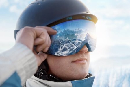 Gros plan des lunettes de ski d'un homme avec le reflet des montagnes enneigées. Une chaîne de montagnes reflétée dans le masque de ski. Portrait d'homme à la station de ski sur fond de montagnes et de ciel
