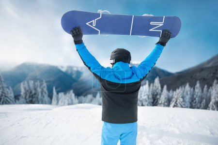 Photo for Snowboarder Of Man At Ski Resort On Blue Sky and Snow Mountains Background, Hold Snowboard.  Winter Sports - Royalty Free Image
