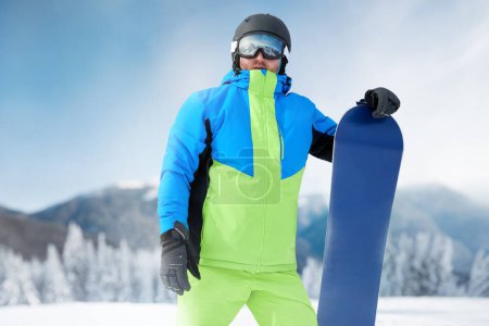 Photo for Snowboarder Of Man At Ski Resort On The Background Blue Sky, Hold Snowboard. Wearing Ski Glasses. Ski Goggles With The Reflection Of Snowed Mountains - Royalty Free Image