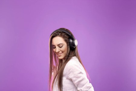 Belle jeune femme dans des écouteurs sans fil écoutant de la musique et dansant sur fond violet. Fille utilise des écouteurs sans fil