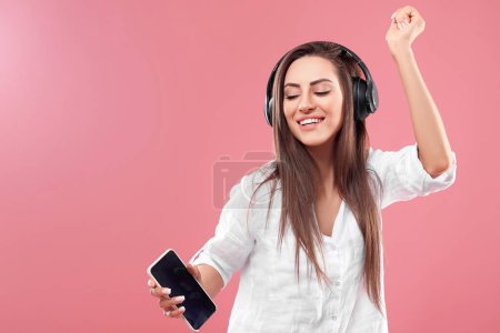 Belle jeune femme dans un casque sans fil écoutant de la musique à l'aide d'un téléphone portable et dansant sur fond rose. Fille utilise des écouteurs sans fil
