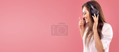 Porträt einer lächelnden brünetten Frau mit Kopfhörer und Smartphone, das Musik über rosa Hintergrund hört. Mädchen benutzt drahtlose Kopfhörer.