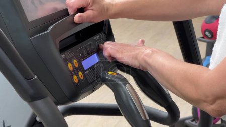 An elliptical cross-trainer is programmed for cardio training by a womans hand. Hands of an elderly woman on the exercise machine. Horizontal video of orbitrek setup on a blurred background of the