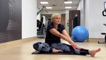 Glückliche Seniorin beim Stretching nach dem Training. 50-60 Jahre alte Kaukasierin in Sportbekleidung sitzt auf dem Boden der Turnhalle.