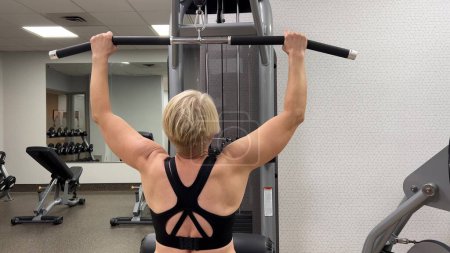 An adult Caucasian woman trains pull-ups with a horizontal bar on a machine in the gym. Rear view, horizontal video. The concept of a sports lifestyle in adulthood. Woman on simulator, back view.