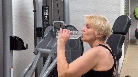 Una mujer utiliza una máquina de fitness y se centra en su entrenamiento. El moderno interior del gimnasio es visible. Una mujer en una máquina de ejercicios bebe agua. Concepto: Lucha por la salud física