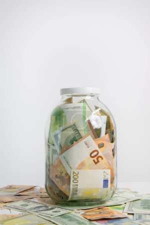 A glass jar filled with US dollars and euro bills sits on a table with scattered bills, ready to be saved or used.