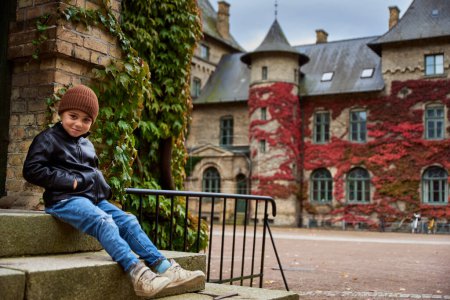 Niño sonriendo por una vieja universidad sueca en un castillo con hiedra roja escalando una pared de ladrillo en otoño. Niño sentado junto a la entrada de un colegio botánico escandinavo transmite el futuro concepto de vida del campus