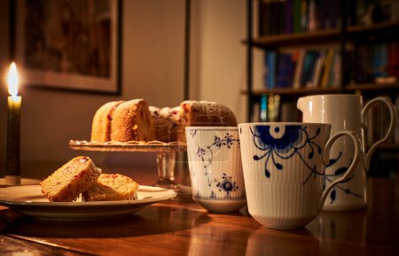 Winter vibe with luxurious mug of hot coffee, doughnut and a library in background, lit by candle light. Donut slice or sliced ciambella in luxury environment lit by burning candle and a book shelf
