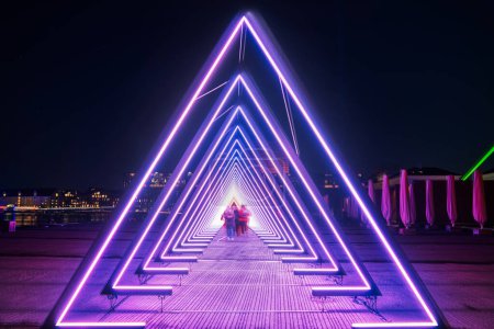 Téléchargez les photos : Porte de couleur pourpre de lumière ou installation de tunnel lumineux violet en néon triangulaire et lumières led la nuit que les gens flous marchent à travers le passage profond lumineux qui ressemble à une trachée humaine Copenhague, Danemark - en image libre de droit