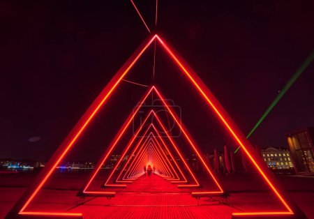 Téléchargez les photos : Série de portes triangulaires de lumière ou tunnel lumineux rouge intense. La porte de l'installation lumineuse se compose d'une succession de portes (triangles) où les gens flous regardent à travers un monde extraterrestre Copenhague, Danemark - en image libre de droit