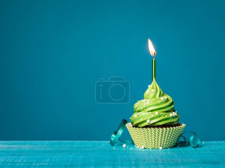 Foto de Magdalena de cumpleaños de crema de mantequilla con glaseado verde, vela encendida, espolvoreos y cinta sobre un fondo azul azulado. - Imagen libre de derechos