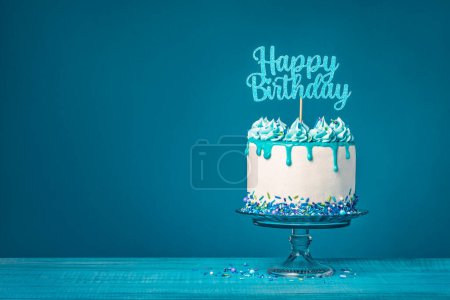 Photo for White drip cake with teal ganache, sprinkles, and a happy birthday topper over a blue background. Simple and trendy. Copy space. - Royalty Free Image
