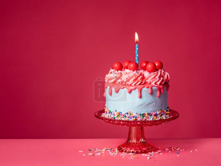 Foto de Bubble Goma de mascar Cumpleaños Drip Cake en un soporte rojo con vela encendida, chispas de colores, glaseado de crema de mantequilla y bolas de caramelo de cereza roja sobre un fondo rosa vibrante. Por encima de la diversión. Copiar espacio. - Imagen libre de derechos