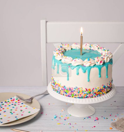 Birthday party place setting with a vanilla buttercream cake, teal blue ganache drip, lit gold candle, and colorful sprinkles on a light grey white background. Copy space.