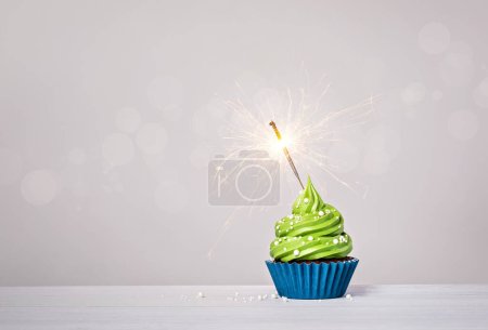 Green birthday cupcake with buttercream icing, blue cup cake liner, sprinkles and lit sparkler on a light grey white background. copy space.