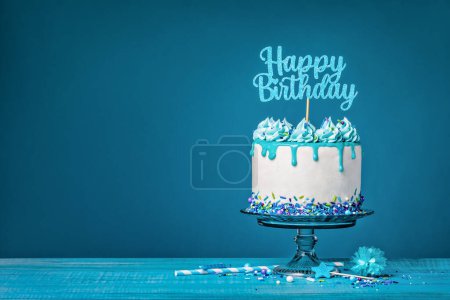 Foto de Pastel de goteo blanco con ganache de teca, salpicaduras y una parte superior feliz cumpleaños sobre un fondo azul. Simple y de moda. Copiar espacio. - Imagen libre de derechos