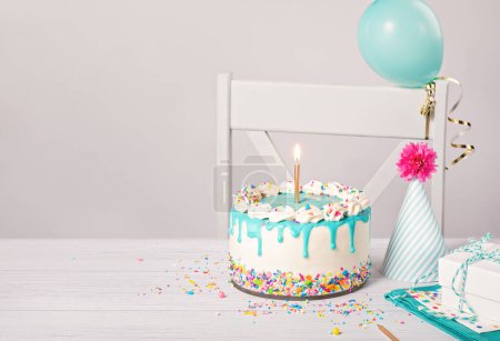 Photo for Birthday party table setting with vanilla buttercream cake, teal blue ganache drip, lit gold candle, and colorful sprinkles on a light grey white background with balloon, hat gifts. Copy space. - Royalty Free Image