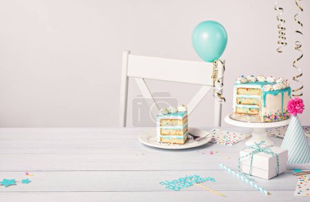 Photo for Happy Birthday party table setting with sliced vanilla confetti cake, teal blue ganache drip on a light grey white background with balloons, ribbon decorations, hat and gifts. Copy space. - Royalty Free Image