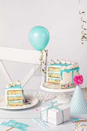 Photo for Happy Birthday party table setting with a sliced vanilla confetti cake, teal blue ganache drip, on light grey white background with balloons, ribbon decorations, hat and gifts. - Royalty Free Image