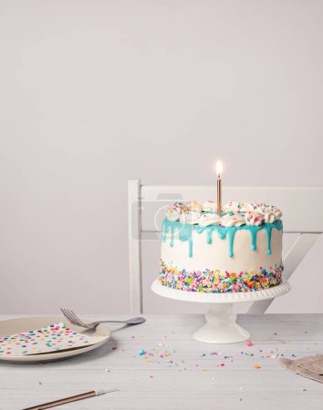Photo for Birthday party place setting with a vanilla buttercream cake, teal blue ganache drip, lit gold candle, and colorful sprinkles on a light grey white background. Copy space. - Royalty Free Image