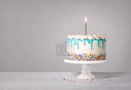 Vanille-Buttercreme-Geburtstagstorte mit kristallblauem Ganache-Tropfen, brennender Goldkerze und bunten Streuseln auf hellgrauem weißem Hintergrund. Kopierraum.