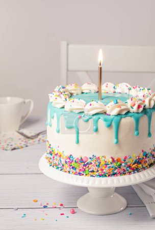 Photo for Close up of a Birthday cake with vanilla buttercream icing, lit gold birthday candle, colorful rainbow confetti sprinkles and teal blue ganache drip on a light grey white table setting background. - Royalty Free Image