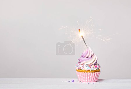 Photo for Purple and pink birthday celebration cupcake with buttercream icing swirl, sprinkles and lit sparkler on a light grey white background. copy space. - Royalty Free Image
