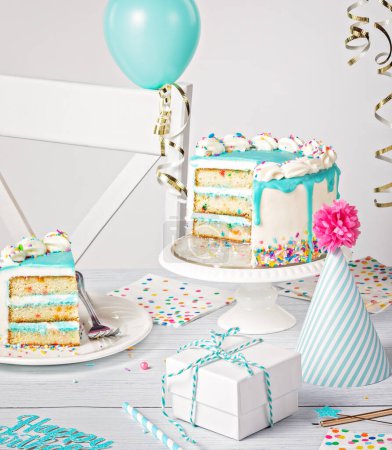 Photo for Birthday party table setting with a teal blue ganache drip cake on a light grey white background with sliced piece of cake and decorations. - Royalty Free Image