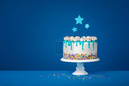 Fun White Birthday cake with trendy teal ganache drips, colorful sprinkles and star toppers over a dark blue background. Copy space.