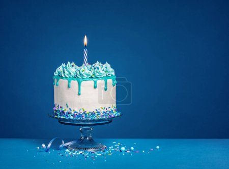 White Birthday drip cake with teal ganache, sprinkles, and a lit candle over a dark blue background. Copy space.