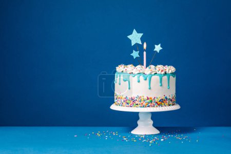 Photo for White Birthday drip cake with three star toppers, a candle and colorful sprinkles over a blue background. Copy space. - Royalty Free Image