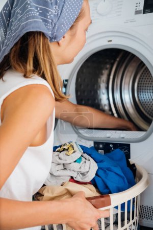 Die junge Haushälterin sitzt vor einer Waschmaschine. Sie lädt Waschmaschine mit schmutziger Wäsche. . Hochwertiges Foto