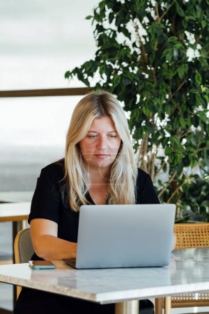 Foto de Mujer rubia joven que trabaja con el ordenador portátil mientras está sentado en la cafetería en el interior. - Imagen libre de derechos