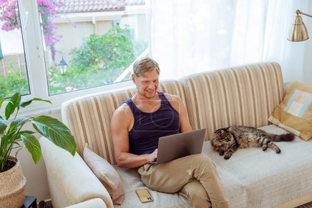 Foto de El hombre se sienta con un portátil cerca del gato en el sofá en casa. Tiempo libre con la tecnología. Foto de alta calidad - Imagen libre de derechos