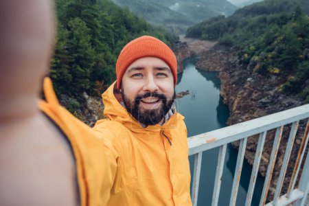 Foto de Caminante tomando retrato de selfie en la cima de la montaña. Chico con sombrero rojo y chaqueta amarilla haciendo reportaje de video o grabando contenido de video para su vlog de viaje - Imagen libre de derechos