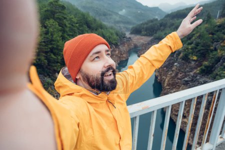 Foto de Caminante tomando retrato de selfie en la cima de la montaña. Chico con sombrero rojo y chaqueta amarilla haciendo reportaje de video o grabando contenido de video para su vlog de viaje - Imagen libre de derechos