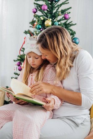 Foto de Madre abrazando a su pequeña hija mientras lee un libro con cuentos de invierno en el sofá en casa junto al árbol de Navidad. - Imagen libre de derechos