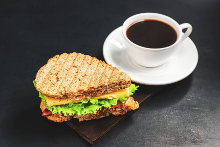 Gegrilltes Sandwich mit Käse, Salat, Schinken und Kaffeetasse auf dunklem Hintergrund. Mittagskonzept mit Kopierraum.