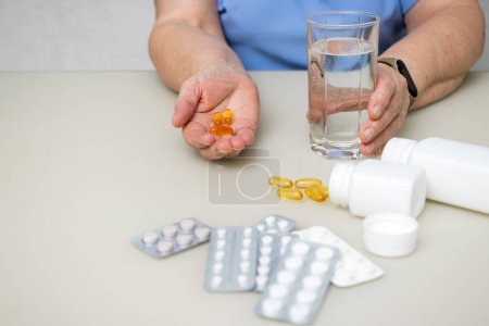 Senior woman with wrinkled old hands at the table holding omega 3 yellow capsules, fish oil pills and water glass. Healthcare and medicine concept.
