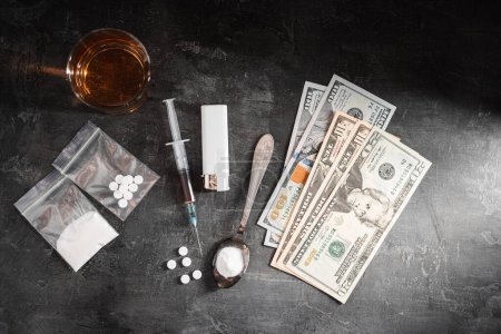 Boisson alcoolisée dans un verre, seringue avec une dose de drogues, pilules blanches dans un sac transparent, poudre de narcotiques dans une cuillère et argent comptant en dollars américains sur fond sombre, vue de dessus.