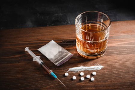Alkoholgetränk im Glas, Spritze mit einer Dosis Drogen, weiße Tabletten und Betäubungspulver in einer durchsichtigen Tüte auf einem Holzbrett. Konzept von Sucht, Missbrauch und schlechten Gewohnheiten.